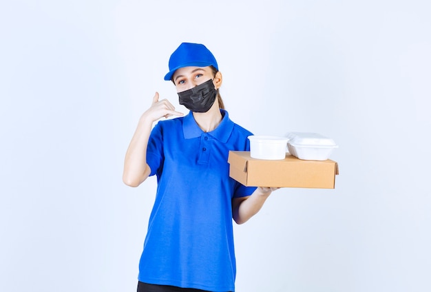 Female courier in mask and blue uniform holding a cardboard box and takeaway packages and asking for a call