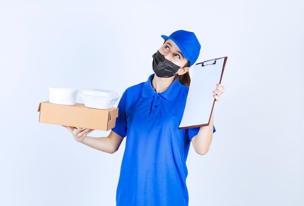 Female courier in mask and blue uniform holding a cardboard box, takeaway packages and presenting the checklist for signature
