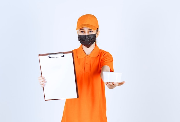 Photo female courier in orange uniform and black mask holding a plastic cup and customer list