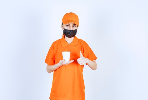 Female courier in orange uniform and black mask holding a takeaway drink and enjoying the taste