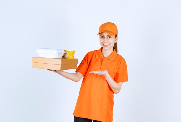 Corriere femminile in uniforme arancione con in mano una scatola di cartone, una scatola di plastica da asporto e una tazza di pasta gialla