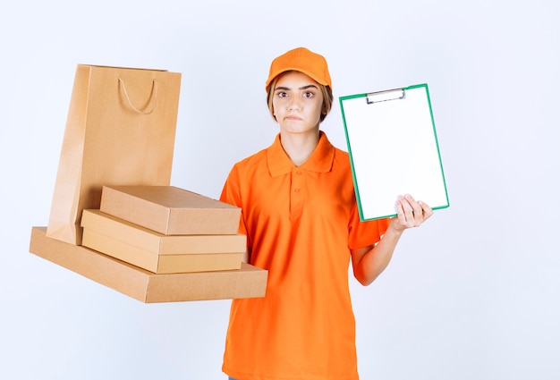 Female courier in orange uniform holding a stock of cardboard parcels and shopping bags, looks confused and presenting the signature list to the customer