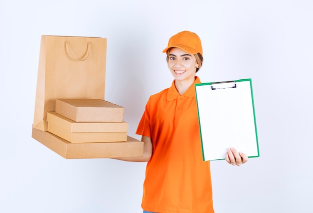 Female courier in orange uniform holding a stock of cardboard parcels and shopping bags and presenting the signature list to the customer