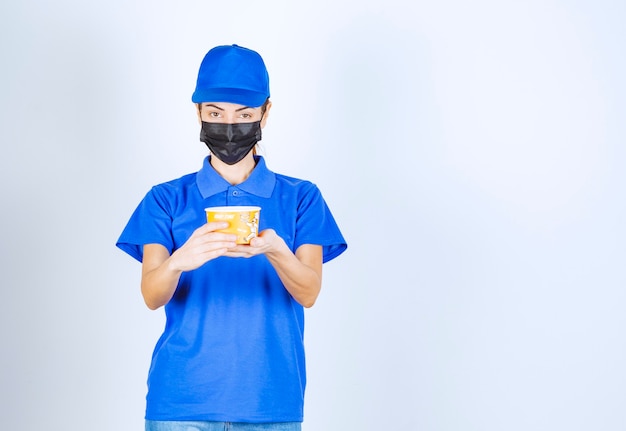 Female courier of the restaurant in blue uniform and face mask holding a takeaway food . 