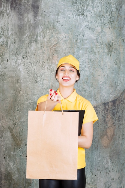 Female courier wearing yellow uniform delivering a cardboard shopping bag and holding a black folder.