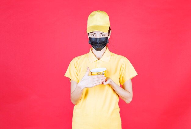 Corriere femminile in uniforme gialla e maschera nera che tiene una tazza di noodles