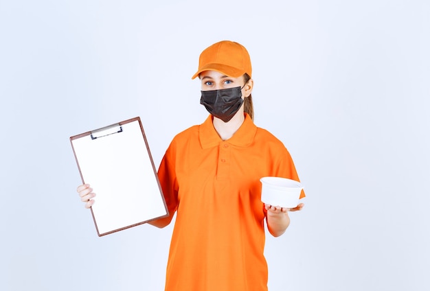 Female courier in yellow uniform and black mask holding a plastic cup and customer list .