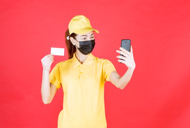 Female courier in yellow uniform and black mask holding a smartphone and taking her selfie while presenting her business card