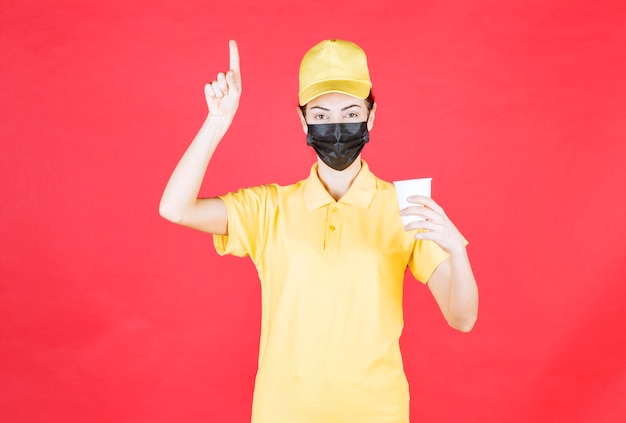 Female courier in yellow uniform and black mask holding a takeaway cup and looks confused and thoughtful