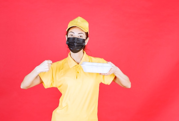 Female courier in yellow uniform and black mask holding a takeaway package and showing her fist