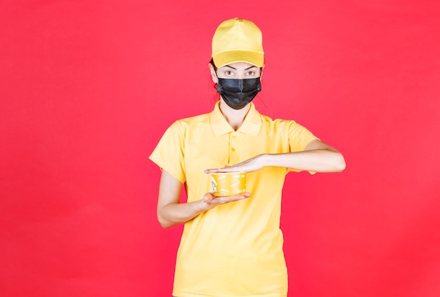 Female courier in yellow uniform and black mask is delivering a noodles cup