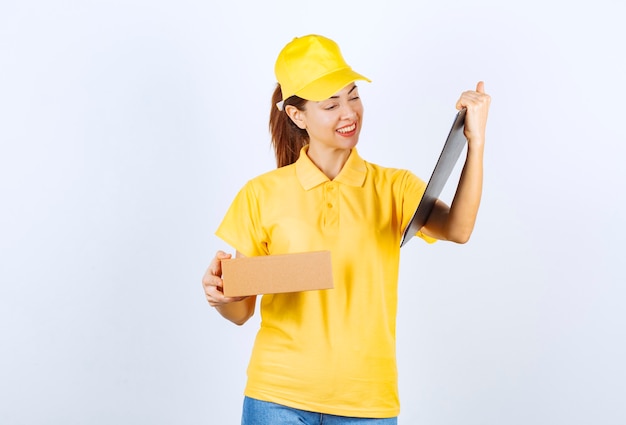 Female courier in yellow uniform delivering the cardboard parcel to the address on the list and looks confused. 