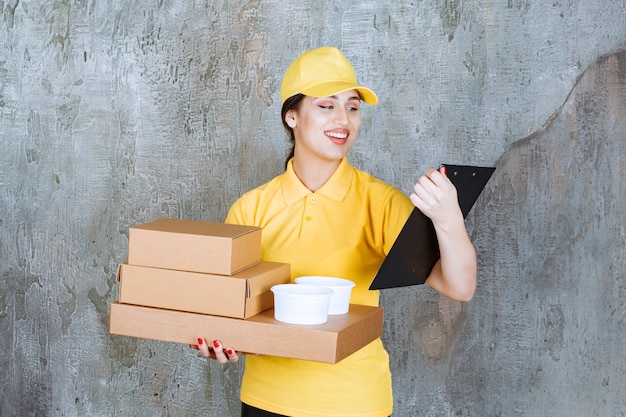 Female courier in yellow uniform delivering multiple cardboard boxes and takeaway cups and checking the address on the list.