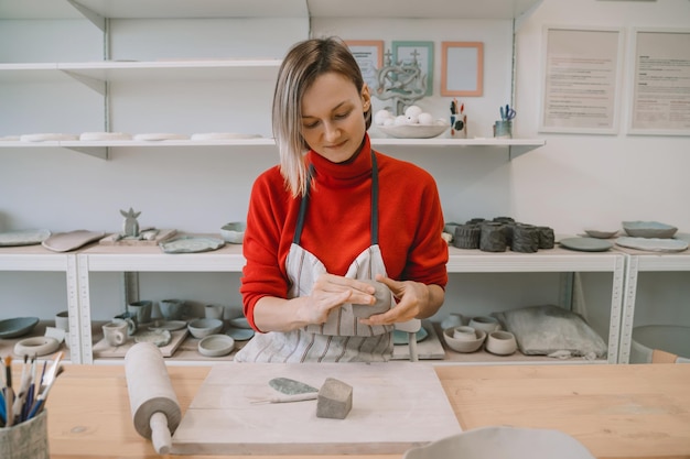 Female craftsperson wearing apron working on ceramics tableware using hands tools on pottery workshop Young woman creating clay products on ceramics workshop Smiling girl in art studio workspace