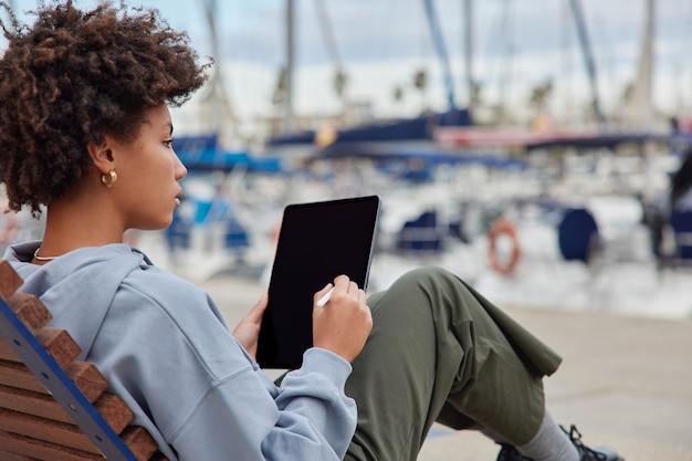 Female creative designer works on digital tablet thinks about creating new project dressed in casual clothes draws on blank screen with stylus poses at pier near harbor uses illustrator application