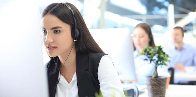 Female customer support operator with headset and smiling, with collegues at background.