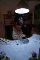 Photo female dark haired goldsmith looking down while sawing jewelry on finagel at table illuminated by lamp