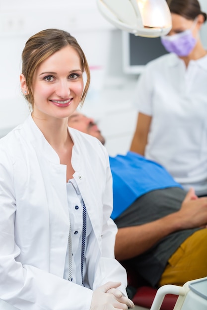 Foto dentista femminile nel suo intervento chirurgico guardando lo spettatore, sullo sfondo la sua assistente sta dando un trattamento a un paziente maschio