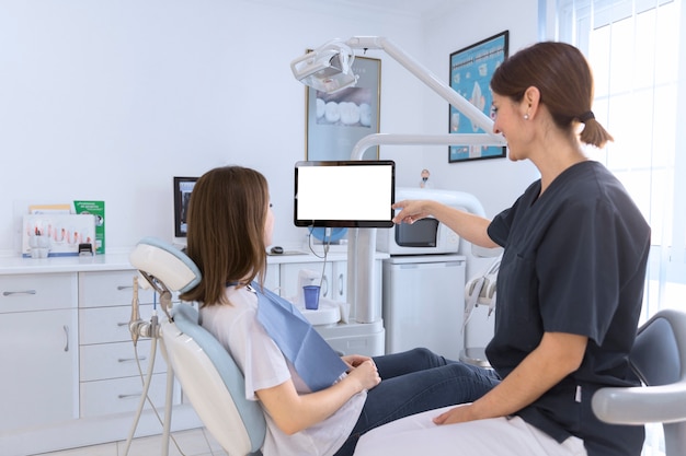 Female dentist pointing on digital screen to female patient in dental clinic