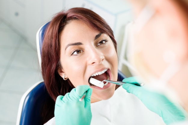 Female dentist preparing patient for dental treatment. She is inserting cotton pad in patient mouth. Selective focus, focus on the patient.