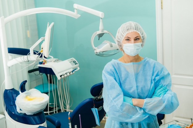 A female dentist wearing a medical mask and rubber gloves poses for the camera and folds her arms in her office.