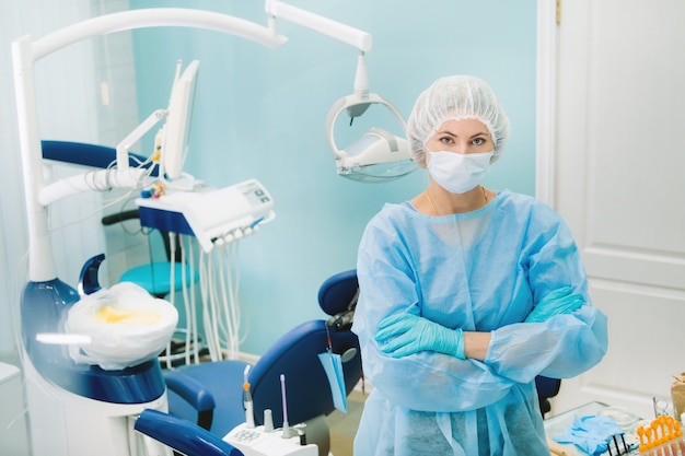 A female dentist wearing a medical mask and rubber gloves poses for the camera and folds her arms in her office