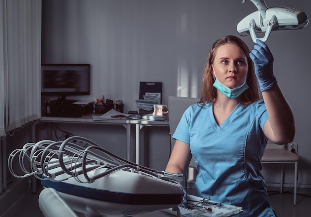 Женщина-стоматолог в униформе и маске регулирует осветительное оборудование над стулом в стоматологической клинике.