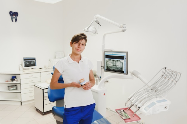 The female dentist with short hair in a white shirt and blue trousers holds dentures in the dental office