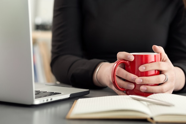 朝のお茶やコーヒーを飲むオフィスの女性デザイナー。勤勉な日のコーヒーブレイク。熱い飲み物のカップを保持している女の子。