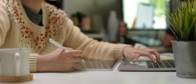 Foto progettista femminile che per mezzo della compressa digitale e scrivendo sul computer portatile sulla scrivania bianca