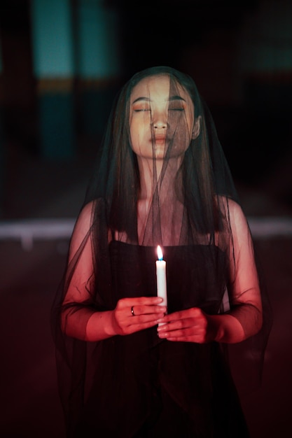 A female devil worshiper with a transparent veil is performing a spooky ritual by holding a candle in her hand