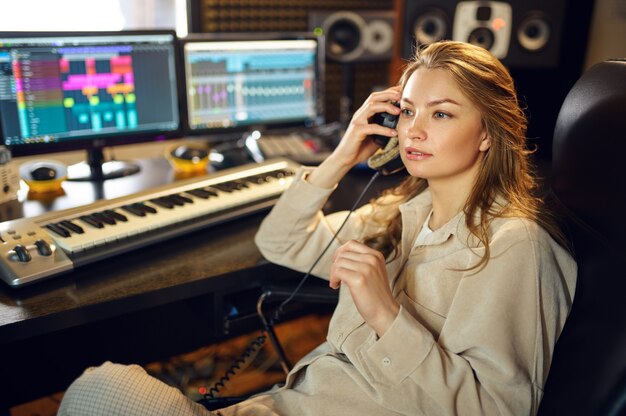 Female dj in headphones listening record, recording studio interior on background. Synthesizer and audio mixer, musician workplace, creative process