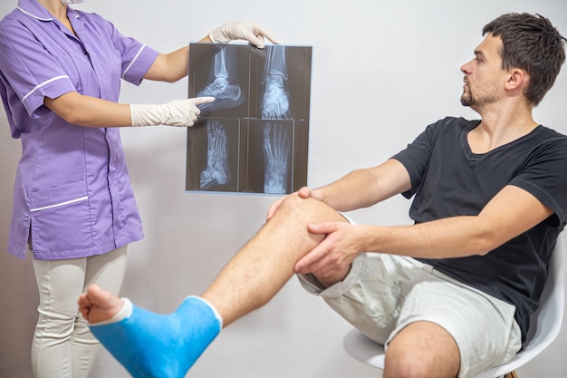 파란색 의료 가운을 입은 여성 의사가 다리가 부러진 남성 환자에게 엑스레이 결과를 설명합니다.
