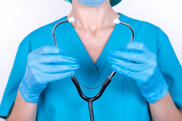 Female doctor in blue uniform and gloves holds a stethoscope in her hands.