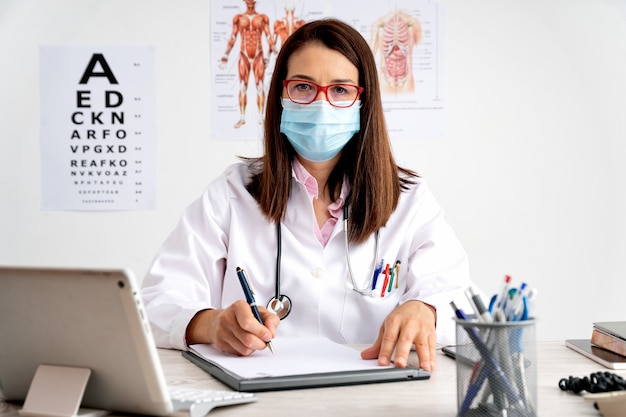 Female doctor in consultation with face mask filling prescription looking at camera