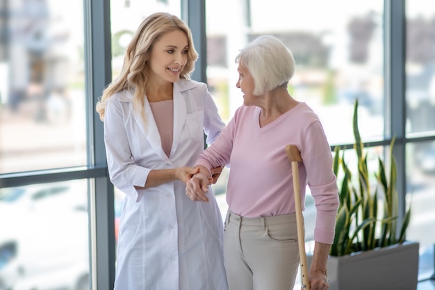 Female doctor and an elderly woman with a crutch walking in a corridor and talking