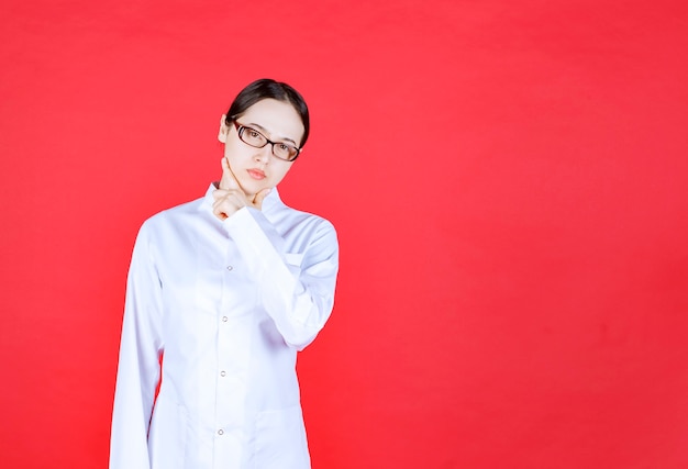 Female doctor in eyeglasses standing on red background, putting hand to her chin and thinking.