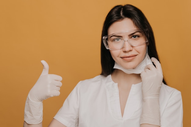 医療ガウンに身を包んだ眼鏡をかけた女医が親指を立て、手術用マスクを脱いだ。