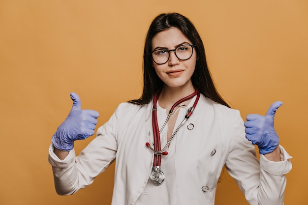 Female doctor in glasses, phonendoscope on her neck dressed in a medical gown, shows thumb up gesture.