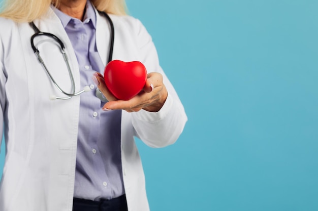 Female doctor in glasses with stethoscope holding heart on the blue background with copy space