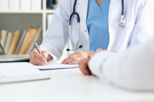 Female doctor hand holds silver pen filling patient list at clipboard pad closeup.