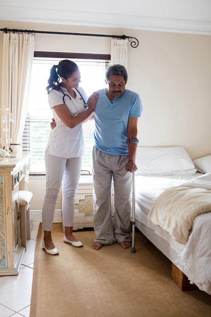 Female doctor helping senior man to walk with walker in bedroom