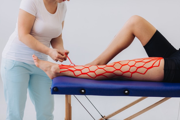 女医は彼女の足にキネシオテープを貼ることによって女性を助けます。彼女の足に運動学弾性治療テープを持つ若い白人女性