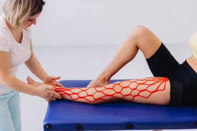 Female doctor helps woman by putting kinesio tape on her leg. Young caucasian woman with kinesiology elastic therapeutic tape on her leg