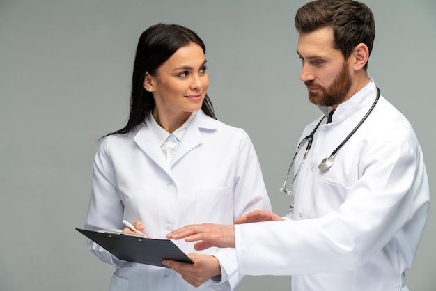 Female doctor holding clipboard and discussing something with her male colleague with stethoscope with concentrated faces while standing isolated on grey background