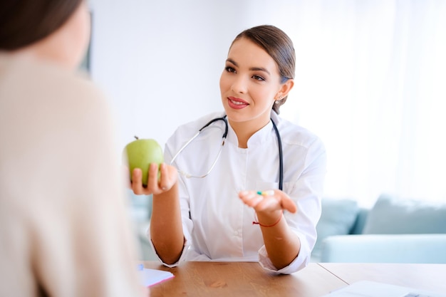 Female doctor holds apple and pills while discussing with patient in the office in hospital