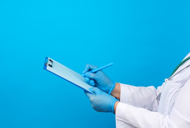 Фото Женщина-врач в белом халате, резиновые синие медицинские перчатки держит папку для бумаг и правой рукой пишет на листе
