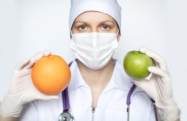 Фото Женский доктор в медицинской защитной маске с концом-вверх плодоовощей. профилактика и здоровое питание во время пандемии.
