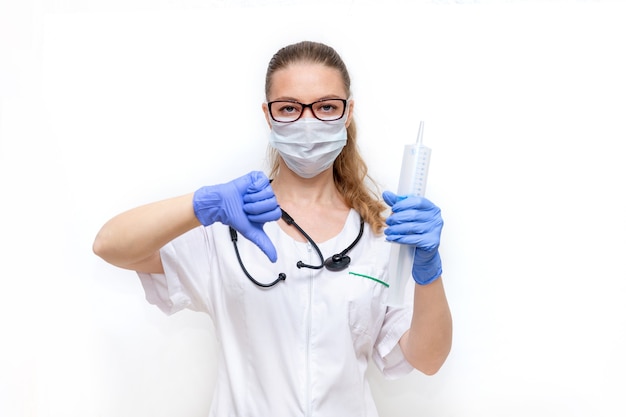 Female doctor in mask holds large syringe and gives thumbs down on white background