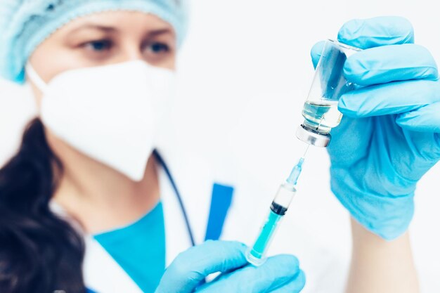 A female doctor in a medical face mask and gloves dials the\
vaccine into a syringe from an ampoule